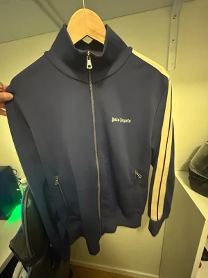 Blå zip-tröja Palm Angels - Snygg blå zip-tröja från Palm Angels med vita ränder längs ärmarna och logga på bröstet. Tröjan har hög krage, dragkedja framtill och två fickor med dragkedja. Perfekt för en sportig och trendig look.