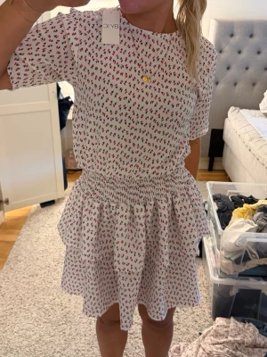 Vit kortklänning med blommigt mönster från BY IC - Supersöt vit kortklänning med små blommor i rött och grönt. Klänningen har korta ärmar, rund halsringning och smock i midjan som ger en fin siluett. Kjolen är volangig och ger en lekfull känsla. Perfekt för varma dagar och sommarhäng.