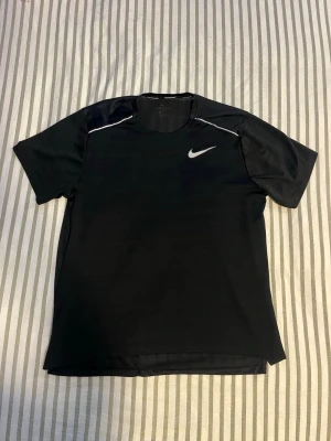 Svart Nike Miler tshirt - Snygg svart tränings-tshirt från Nike med korta ärmar och vit logga på bröstet. T-shirten har vita detaljer vid axlarna och är gjord i ett lätt, snabbtorkande material som passar perfekt till träning och sport.