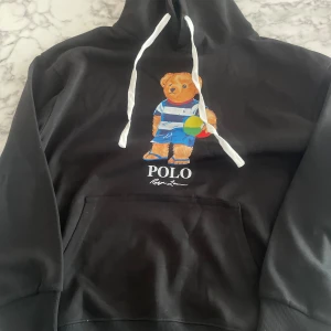 Svart Polo Ralph Lauren hoodie med björn - Svart hoodie från Polo Ralph Lauren med ikonisk björnprint på bröstet. Tröjan har vit snörning i huvan, stor magficka och långa ärmar. Björnen har strandstil med randig tröja och badboll. Perfekt för chill dagar och streetwear. Använd ett par gånger men inga defekter. stolek L