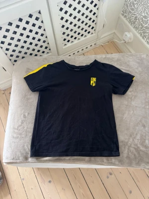 Svart AIK t-shirt med gula detaljer - Säljer en svart t-shirt med AIK-logga på bröstet och texten STOCKHOLM i gult på ryggen. T-shirten har gula ränder på axlarna och är kortärmad. Perfekt för dig som vill representera AIK och Stockholm. Denna T-shirt är köpt på norra från ultras nord och går ej att få tag på längre.