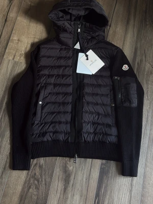 Svart pufferjacka med huva Moncler - Snygg svart pufferjacka från Moncler med stickade ärmar och quiltad front. Jackan har huva, dragkedja framtill och fickor med tryckknappar. Moncler-logga på ärmen och en extra ficka med dragkedja på vänster arm. Perfekt för kyliga dagar.