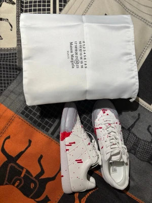 Maison Margiela vita sneakers med röd färg - Maison Margiela vita sneakers i läder med unika röda färgstänk över hela skon. Klassisk låg modell med vita snören och gummisula. Snygg och konstnärlig design som sticker ut och passar perfekt till streetwear.