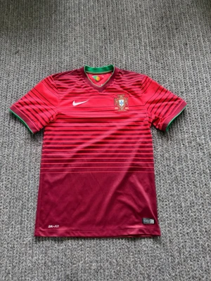 Portugal Nike fotbollströja röd - Säljer en röd Portugal fotbollströja från Nike med svarta horisontella ränder, gröna detaljer vid krage och ärmslut samt Portugal-emblem på bröstet. Tröjan har korta ärmar och är tillverkad i lätt, snabbtorkande Dri-FIT material.