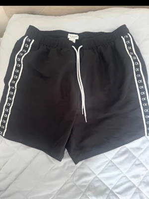 Svarta badbyxor från Calvin Klein - Svarta badbyxor från Calvin Klein med vita snören i midjan och vita sidoremsor med CK-logga längs båda benen. Elastisk midja för skön passform. 
