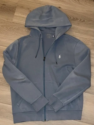 Ljusblå Ralph Lauren zip hoodie - Säljer denna stilrena zip hoodie från Polo Ralph Lauren i en ljusblå nyans. Hel dragkedja, huva med dragsnören och klassisk broderad logotyp på bröstet. Mjuk och bekväm modell som passar både till vardags och fritid. I fint skick. Nypris 1400-2000