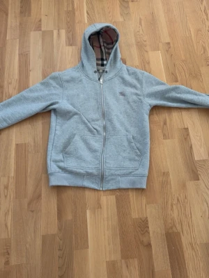 Grå hoodie med dragkedja och huva - Säljer en grå hoodie med dragkedja och huva. Insidan har det klassiska rutiga mönstret i beige, svart och rött. Två fickor framtill och ribbade muddar vid ärmslut och nederkant. Perfekt för en chill och stilren look.
