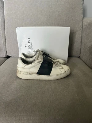 Valentino open skor - Snygga vita sneakers från Valentino med en bred svart läderdetalj över snörningen. Skorna har en chunky sula och nitar längs hälen för en cool touch. Tillverkade i läder och har en klassisk låg profil som passar till många olika stilar.
