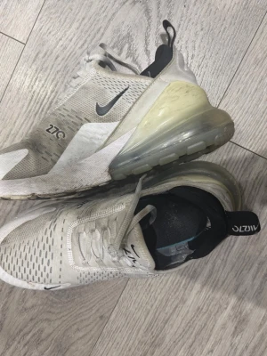 Nike Air Max 270 vita sneakers - Nike Air Max 270 sneakers i vitt med svarta detaljer och stor synlig luftbubbla i hälen. Ovandelen är i mesh för bra ventilation och har en modern, sportig look. Swoosh-logga på sidan och 270-tryck. Skön passform och snygg design.