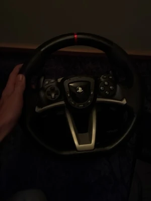Ratt och pedaler till PlayStation och PC - Säljer en svart ratt med PlayStation-logga och tillhörande pedaler, perfekt för racing- och bilspel. Ratten har flera knappar och en klassisk rund form, tillverkad i plast och gummi för bra grepp. Pedalerna är också i svart plast och kopplas enkelt till konsolen. Varan har andvände fåtal kanske 3-5 gånger
