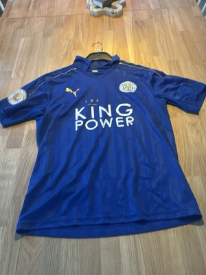 Blå Leicester City matchtröja Puma - Säljer en blå Leicester City matchtröja från Puma med klubbmärke och King Power-tryck på bröstet. Tröjan har korta ärmar, guldfärgade detaljer och nummer 20 på ryggen. Perfekt för dig som älskar fotboll och vill visa ditt stöd för laget.