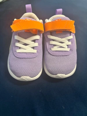 Lila sneakers med orange kardborre st 23 barn  - Snygga sneakers Puma  i lila mesh med vita snören och en cool orange kardborre. Skorna har vit sula och detaljer i lila och vitt. Perfekta för dig som gillar färg och vill sticka ut lite extra. Lätta att ta på tack vare kardborrebandet.
