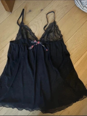 Svart spetsnattlinne med rosett - Sött svart nattlinne med tunna axelband och spetsdetaljer vid bysten. Dekorerad med en liten rosett i blommigt tyg framtill. Materialet är tunt och lätt, perfekt för mysiga kvällar hemma.