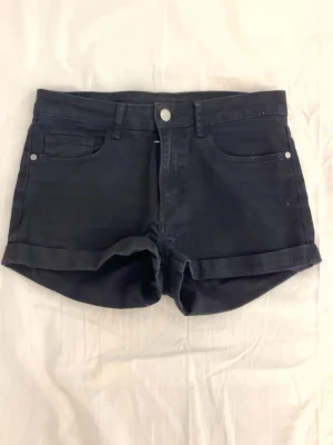 Svarta lågmidjade denimshorts med upprullad kant - Snygga svarta shorts i denim med upprullad kant och klassisk femficksdesign. De har normal passform och bälteshällor i midjan. Perfekta för varma dagar och enkla att matcha med olika stilar.