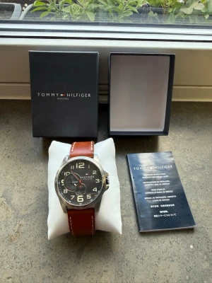 Tommy Hilfiger klocka med brunt läderband - Klockan har en tidlös design som passar lika bra till vardags som till mer formella tillfällen. Den har en robust boett i rostfritt stål, en stilren svart urtavla med tydliga siffror och snygga röda detaljer. Det bruna läderarmbandet har vita kontrastsömmar som ger en exklusiv känsla. Klockan säljes komplett i sin originalbox tillsammans med den officiella instruktionsboken och garantiguiden. OBS Batteri behöver bytas! 