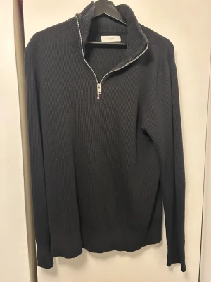 Svart stickad tröja med halv dragkedja - En svart stickad tröja med hög krage och halv dragkedja framtill. Tröjan har lång ärm och är ribbstickad för en snygg och enkel look. Perfekt för lager på lager och passar till det mesta i garderoben.
