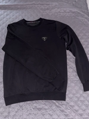 Svart sweatshirt från Prada - Snygg svart sweatshirt från Prada med klassisk rund hals och lång ärm. Tröjan har en ikonisk triangel-logga på bröstet som ger en exklusiv känsla. Perfekt för dig som gillar stilrena och lyxiga plagg.