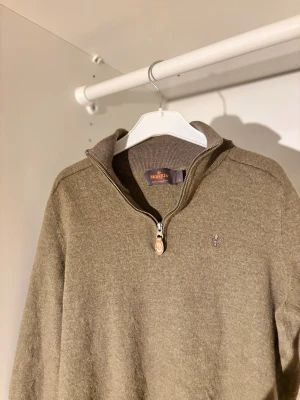 Olivgrön halvzip tröja Morris - Olivgrön stickad tröja från Morris med halvzip och hög krage. Tröjan har ribbade muddar vid ärmslut och nederkant samt en liten broderad logga på bröstet. Perfekt för lager på lager och snygg till jeans eller chinos. Storleken är small i barn storlek och den passar uppskattningsvis på 140-150cm lång och den har en liten defekt på sidan som i princip är osynlig