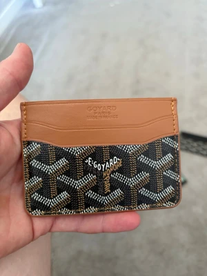 Snygg korthållare  - Snygg korthållare med klassiskt mönster i svart, brunt och vitt. Framsidan har  Paris-logga och flera kortfack. Övre delen är i ljusbrunt läder och resten i canvas med  ikoniska tryck. Perfekt för dig som vill ha stilren och lyxig accessoar.