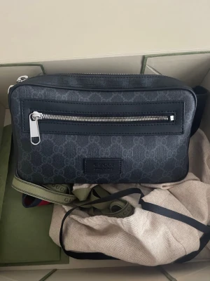 Äkta gucci belt bag - Snygg svart äkta axelväska från Gucci med diskret GG-mönster, silverfärgade dragkedjor och justerbar axelrem med röd-svarta detaljer. Väskan är tillverkad i canvas och läder, har ett ytterfack med dragkedja och stilren design. Obs kvitto eller box medföljer inte.