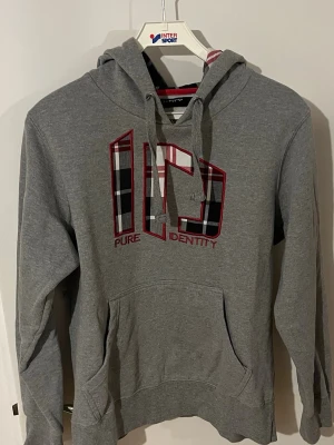 Grå hoodie med rutigt tryck - Snygg grå hoodie med stor ficka framtill och huva med rutigt innerfoder. Framsidan har ett stort tryck med texten 'Pure Identity' och rutigt mönster i svart, vitt och rött. Perfekt för en avslappnad och trendig stil. Pris kan diskuteras vid snabb affär
