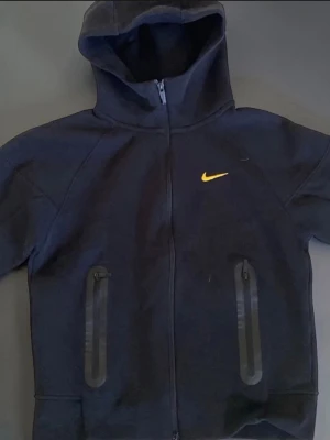 Svart Nike hoodie med gul logga - Snygg svart hoodie från Nike med dragkedja och huva. Framtill finns två fickor med svarta dragkedjor och en gul Nike-logga på bröstet. På baksidan syns små gula broderade detaljer. Perfekt för en sportig och avslappnad stil.