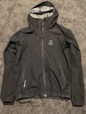 Svart regnjacka från Haglöfs - Snygg svart regnjacka från Haglöfs med huva och hel dragkedja framtill. Jackan har två dragkedjeförsedda fickor och är tillverkad i vattentätt material med Gore-Tex-teknologi. Perfekt för regniga dagar och outdoor-äventyr.