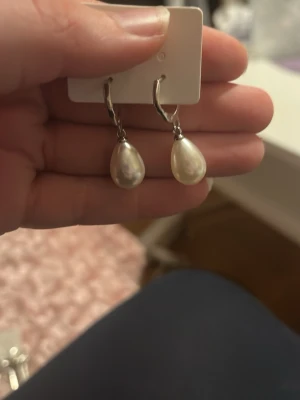 Silvriga örhängen med droppformade pärlor - Snygga örhängen i silverfärg med droppformade vita pärlor som hänger från små ringar. Perfekta för dig som gillar klassiska och stilrena accessoarer med en elegant touch.