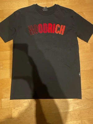 Svart HOODRICH t-shirt med röd text - Svart t-shirt från HOODRICH i storlek S. Framtill står det HOODRICH i stora bokstäver med en snygg färgskiftning från rött till orange. Klassisk rund hals och korta ärmar. Perfekt för dig som gillar streetwear och vill sticka ut.