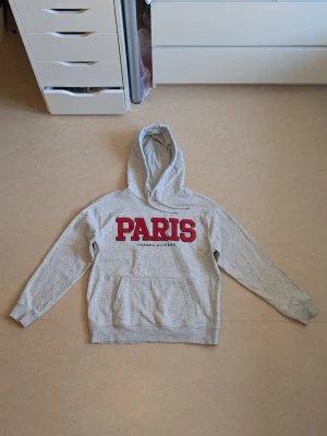 Grå hoodie med PARIS-tryck - HELT OANVÄND HOODIE. Snygg grå hoodie med stor röd PARIS-text på bröstet och mindre svart text under. Klassisk huva och magficka framtill. Perfekt för en avslappnad streetwear-look. Tillverkad i mjukt sweatshirtmaterial och har ribbade muddar vid ärmar och nederkant. Jag har tvättat hoodie 1 gång maskintvätt precis innan jag la upp den för försäljning på Plick