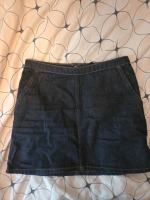 Mörkblå jeanskjol med dragkedja - Snygg mörkblå jeanskjol i kort modell med framfickor och synliga sömmar. Kjolen har en dragkedja och knapp framtill för en klassisk denimlook. Perfekt att matcha med en enkel t-shirt eller stickad tröja.