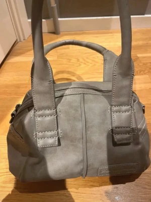 Grå handväska med breda handtag - Snygg grå handväska i mjukt material med breda, dubbla handtag och dekorativa sömmar. Väskan har en rundad form och dragkedja upptill. Perfekt för dig som vill ha en stilren och enkel accessoar till din outfit.