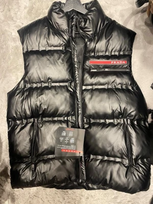 Svart dunväst från Prada - Snygg svart dunväst från Prada med hög krage och dragkedja framtill. Västen har två sidofickor med dragkedja och en diskret logga på bröstet. Tillverkad i glansigt material som ger en exklusiv känsla. Perfekt för lager-på-lager och streetwear.