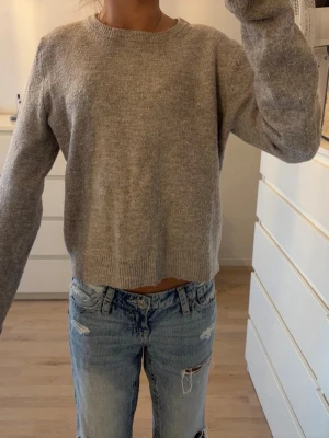 Grå stickad tröja från H&M - Mysig grå stickad tröja från H&M med rund halsringning och långa ärmar. Tröjan har en enkel, rak passform och ribbade muddar vid ärmslut och nederkant.
