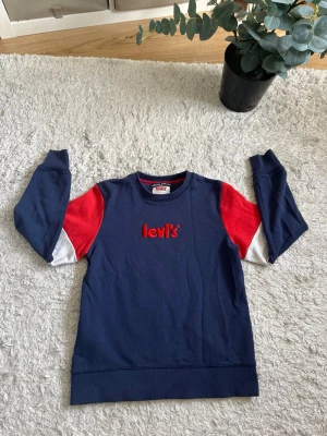 Blå Levi's sweatshirt - Snygg sweatshirt från Levi's i marinblått med röda och vita detaljer på ärmarna. Tröjan har rund halsringning, långa ärmar och Levi's-logga broderad i rött framtill. Perfekt för en avslappnad och sportig stil. Om du har några frågor skriv till mig, svarar så snabbt jag kan.🙌