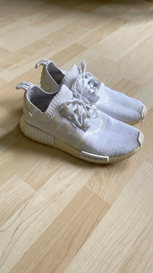 Adidas NMD R1 Primeknit Japan - Säljer ett par vita Adidas NMD R1 sneakers med primeknit tyg och klassiska tre ränder på sidan. Skorna har vita snören, textdetaljer på sidan och bak, samt en bekväm Boost-sula.