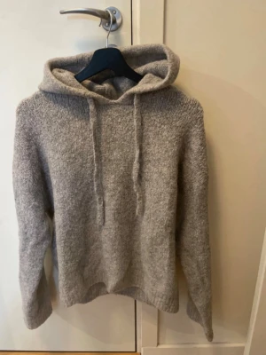 Mysig grå stickad hoodie - Säljer en supermjuk och varm grå stickad hoodie med huva och dragsnören. Tröjan har en avslappnad passform, långa ärmar och ribbade muddar. Perfekt för dig som gillar en enkel men stilren look. Materialet känns fluffigt och bekvämt.
