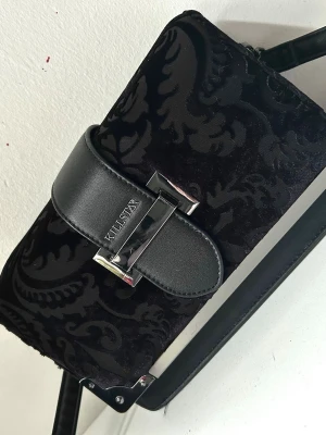 Svart handväska från Killstar - Snygg svart handväska från Killstar med sammetsliknande material och barockinspirerat mönster. Väskan har ett brett lock med metallspänne och logotyp, samt detaljer i silver. Axelremmen är justerbar och väskan har en rektangulär form.