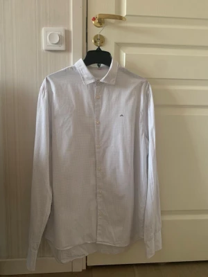 J Lindeberg Skjorta - Klassisk vit J.Lindeberg skjorta med diskret rutmönster och knäppning framtill. Skjortan har lång ärm, krage och en liten broderad detalj på bröstet. Perfekt för en clean och stilren look. Skick 9.5/10 och storlek S passar dig runt 170cm-178cm