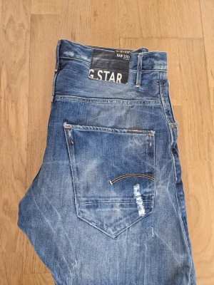 Blå jeans från G-Star RAW - Säljer ett par blå jeans från G-Star RAW med slitna detaljer. Jeansen har en klassisk femficksmodell och tydliga tvättade effekter för en cool, avslappnad look. Materialet är denim i bomull och passformen är rak. Storlek: W:34, L:34. Köpta från vinted