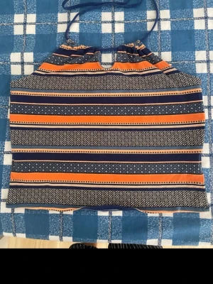 Randig halterneck linnetopp - Säljer en färgglad halterneck linnetopp med randigt mönster i orange, blått och vitt. Toppen har tunna band som knyts i nacken och ett coolt grafiskt mönster mellan ränderna. Perfekt för varma dagar och ger en avslappnad vibe.