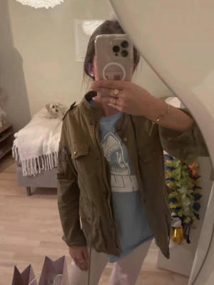 Olivgrön parkajacka med fickor - Säljer en olivgrön parkajacka med flera fickor och axeldetaljer. Jackan har knappar framtill och patch på ena ärmen. Perfekt för dig som vill ha en avslappnad och cool stil.