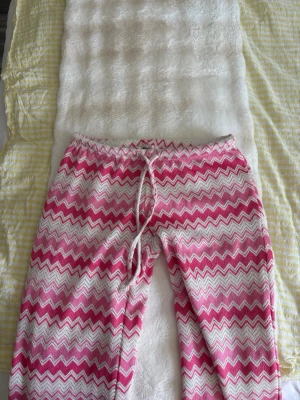 Rosa och vita zigzag-mönstrade byxor - Säljer ett par stickade byxor med rosa och vita zigzag-ränder. De har dragsko i midjan och är gjorda i ett mjukt, stretchigt material. Perfekta för dig som gillar färg och mönster i din outfit. De är extremt eftertraktade och lågmidjade!