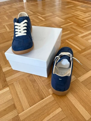   Maison Margiela gats storlek 43 - Snygga blå sneakers från Maison Margiela med vita skosnören och beige gummisula. Skorna har en mix av mocka och läderdetaljer, klassisk låg silhuett och diskret logga på plösen. Perfekta för dig som gillar stilrena och trendiga sneakers.