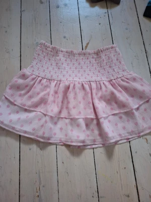 Rosa kortkjol från Gina Tricot - Söt rosa kortkjol från Gina Tricot med volang och smock i midjan. Kjolen har ett diskret mönster i ljusrosa och är gjord i ett lätt och luftigt material, perfekt för varma dagar.