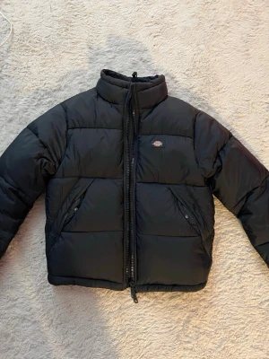 Svart pufferjacka från Dickies XS - Säljer en svart pufferjacka från Dickies i storlek XS. Jackan har hög krage, två snedställda fickor med knappar och dragkedja framtill. Klassisk Dickies-logga på bröstet och vadderad insida för extra värme. Perfekt för kalla vinterdagar.