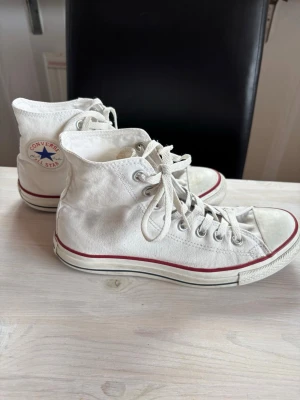 Vita Converse Chuck Taylor Highs - Klassiska vita Converse Chuck Taylor All Star high tops med röd rand runt sulan och vita skosnören. Skorna har canvasmaterial, metallöljetter och den ikoniska loggan på sidan. Perfekta för en avslappnad och tidlös stil.