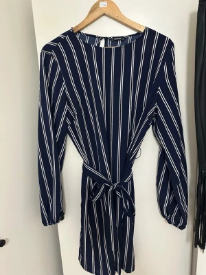 Mörkblå randig jumpsuit från SHEIN - Snygg mörkblå jumpsuit från SHEIN med vita vertikala ränder. Modellen har långa ärmar med resår vid handlederna, rund halsringning och ett knytband i midjan för en markerad siluett. Perfekt för dig som gillar stilrena och trendiga plagg.
