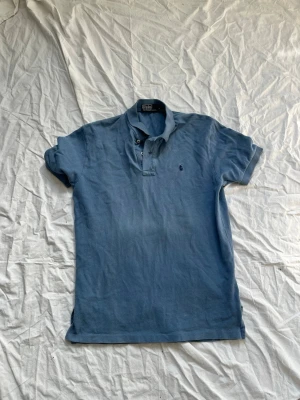 Blå pikétröja från Ralph Lauren - Klassisk blå pikétröja från Ralph Lauren med korta ärmar och krage. Tröjan har två knappar vid halsen och den ikoniska broderade loggan på bröstet. Perfekt till en avslappnad men ändå stilren look.
