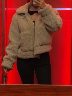 Beige teddyjacka med krage - Mysig beige teddyjacka med stor krage och spänndetalj vid halsen. Jackan har två stora fickor framtill och dragkedja. Boxig och croppad modell som ger en trendig look. Perfekt för kalla dagar när du vill vara både varm och snygg.